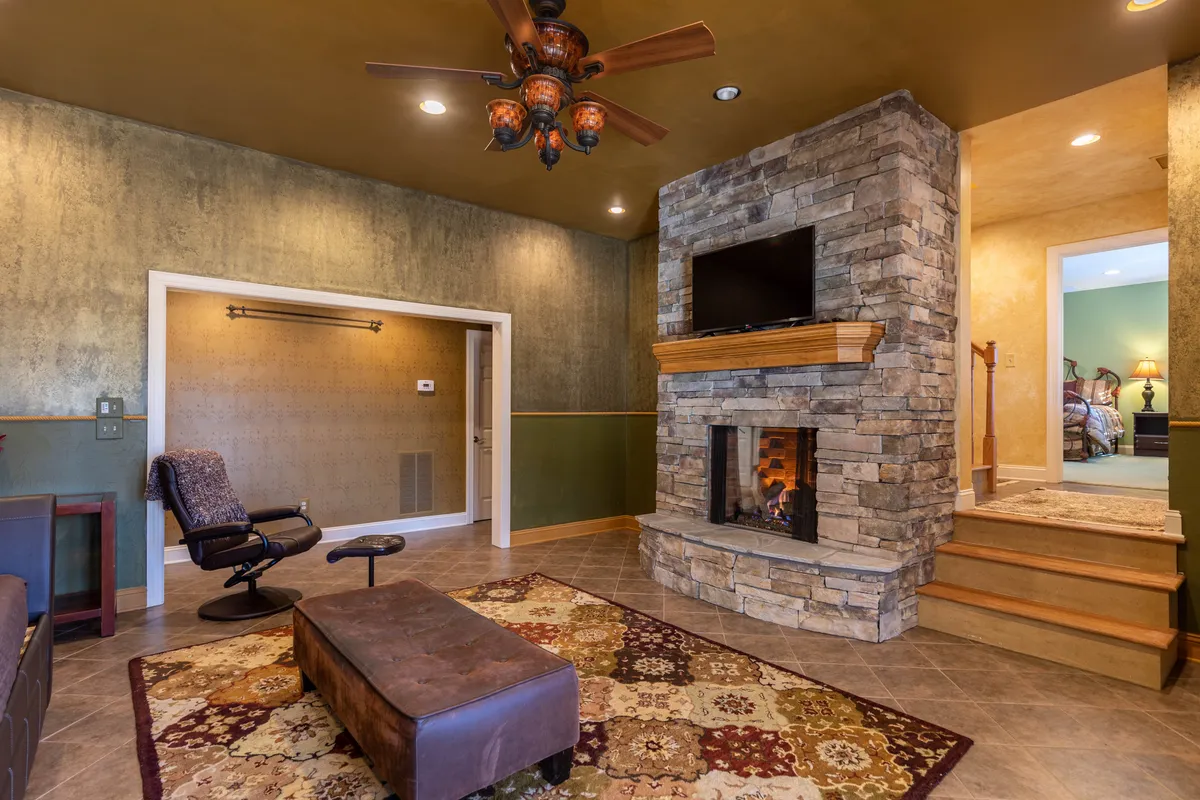 This inviting den features a prominent stone fireplace with a TV mounted above, a cozy colorful area rug, and wooden steps leading to another room. Comfortable seating arrangements create an ideal setting for relaxing, watching movies, or gathering with loved ones for an evening indoors.