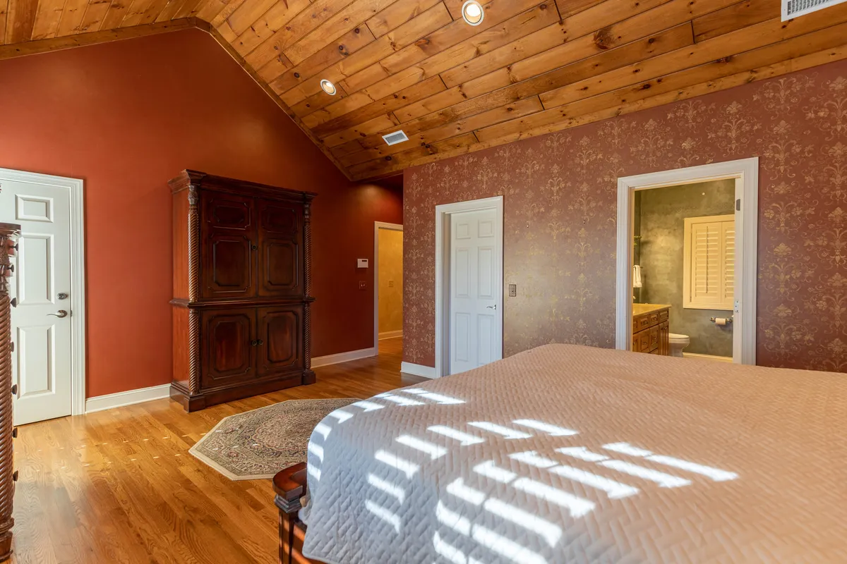 A comfortable bedroom with a wooden ceiling, ornate armoire, a bed with a decorative quilt, and a cozy rug. The inviting space is bathed in soft, natural light, creating a warm environment ideal for unwinding and relaxation.