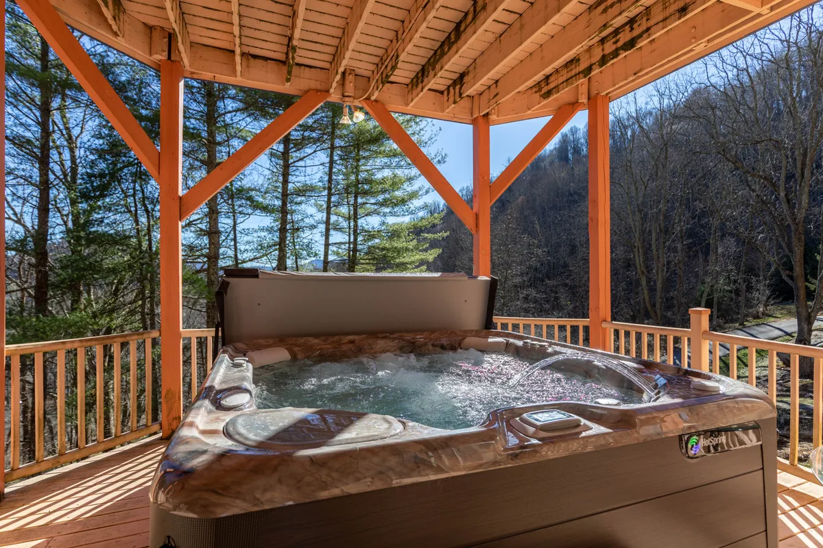 A wooden balcony nestled among trees, featuring a bubbling hot tub with steam rising and comfortable seating nearby. Surrounded by nature, the setting is intimate and inviting, ideal for relaxation and sunset views.