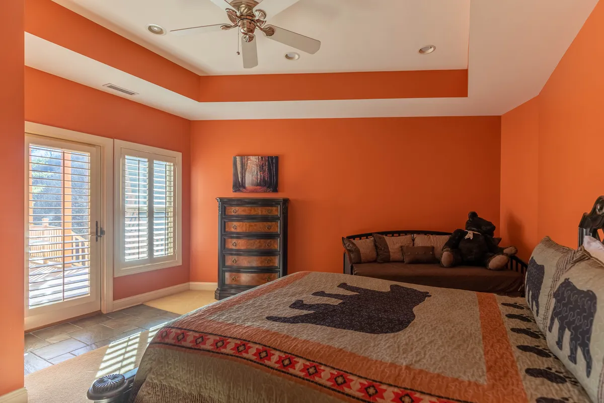 A cozy, warmly-lit bedroom with orange walls, a bear-patterned quilt, and large windows. The room feels inviting and comfortable, offering both a restful spot and a place to share quiet conversations. Natural light pours in, enhancing the sense of retreat and privacy.