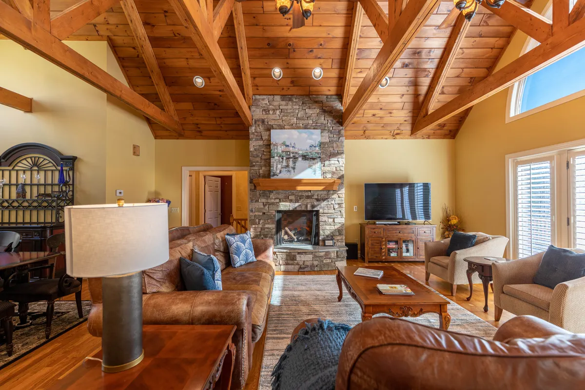 A spacious Great Room featuring soaring wooden beams overhead, a dramatic stone fireplace as the centerpiece, and plush seating arranged for group conversation or movie nights. The inviting mood is set by warm lighting and tasteful décor, making the space ideal for gatherings.