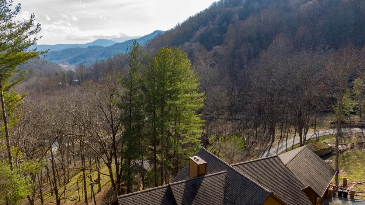 A breathtaking view of a mountainous landscape framed by lush trees, with a winding road visible and the roof of the house peeking out below. The scene offers a sense of peaceful seclusion and natural beauty, ideal for a romantic or luxurious getaway.