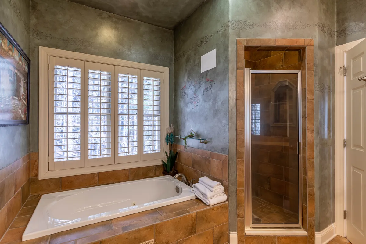 A spa-inspired bathroom featuring a deep soaking tub, a separate walk-in shower, a window with elegant shutters, and textured walls. The setup exudes tranquility and luxury, inviting guests to unwind after a long day.