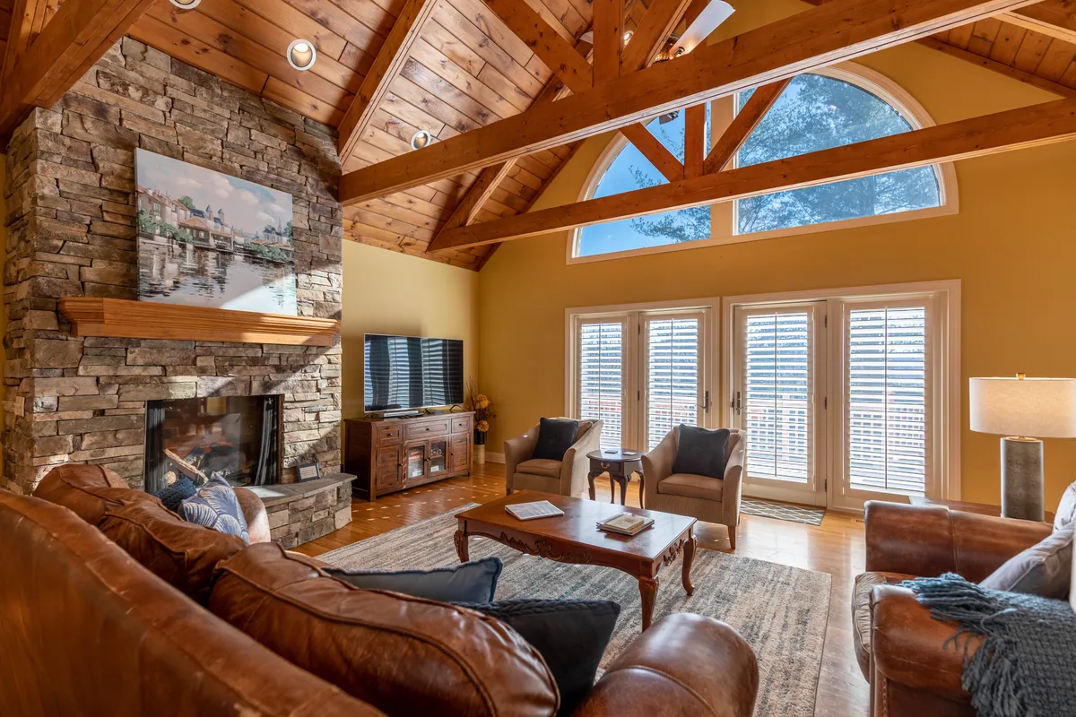 The image showcases a spacious Great Room with wood-beamed vaulted ceilings, a striking stone fireplace, and large windows flooding the room with natural light. Plush, comfortable seating invites guests to gather and relax while taking in the scenic views.